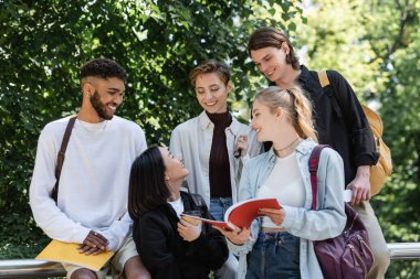 Asian student with takeaway drink talking to friends with notebooks in park 