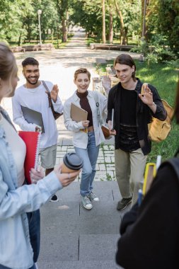 Positive interracial students with gadgets waving hands at friends in park 