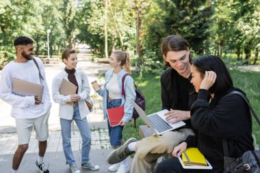 Smiling student holding laptop near asian friend in summer park 