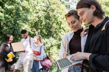 Smiling students using laptop near blurred interracial friends in park 