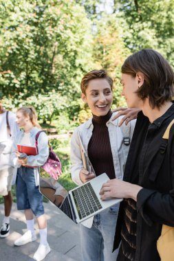 Positive students with laptop talking near blurred multiethnic friends in park 