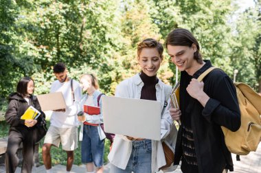 Cheerful students using laptop near blurred multicultural friends in park 