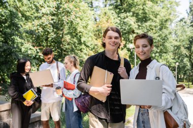 Smiling students with laptop looking at camera near blurred multiethnic friends in park 