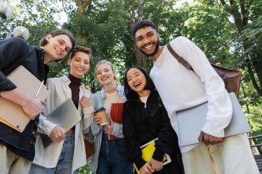 Low angle view of cheerful multicultural students with notebooks and laptops looking at camera in park 