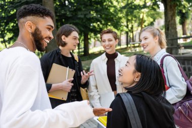 Smiling interracial students talking near blurred friends with backpacks in summer park 