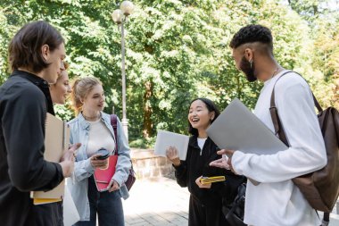 Asian student holding digital tablet near smiling multiethnic friends in summer park 