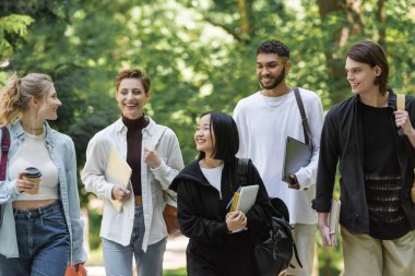Cheerful interracial students with laptops walking in blurred park 