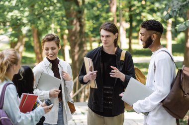 Positive interracial students with laptops talking to blurred friends in park 