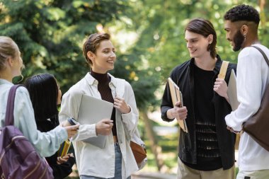 Positive students holding laptops and talking near interracial friends in summer park 