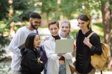 Smiling student holding laptop near interracial friends in summer park 