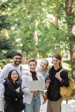 Multiethnic students holding laptop and smiling at camera in park 