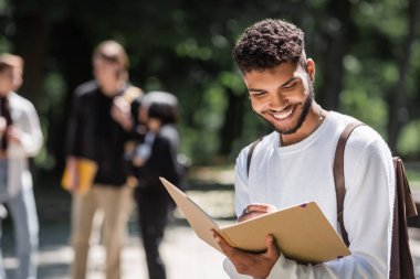 Smiling african american student with backpack writing on notebook outdoors 