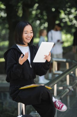 Smiling asian student having video call on digital tablet in park 