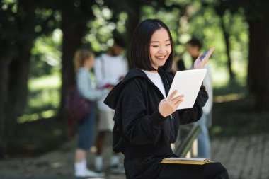Positive asian student having video call on digital tablet in park 