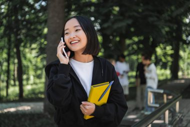 Pozitif Asyalı öğrenci kitap tutuyor ve parkta akıllı telefondan konuşuyor. 