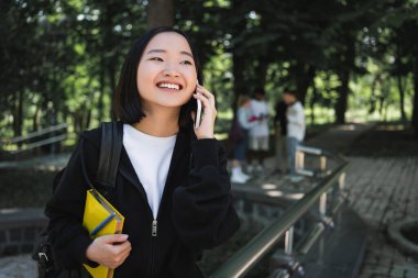 Kitaplı ve sırt çantalı mutlu Asyalı öğrenci parkta akıllı telefonla konuşuyor. 