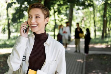Bulanık parkta sırt çantalı olumlu bir öğrenci cep telefonuyla konuşuyor. 