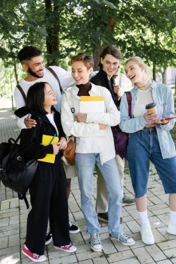 Cheerful multicultural students with copy books and backpacks spending time in park 