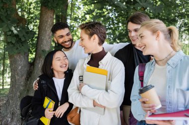 Cheerful multicultural students with copy books talking in park 