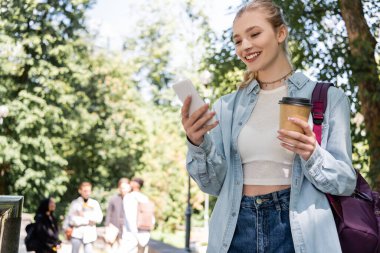 Akıllı telefon kullanan ve parka gitmek için kahve tutan gülümseyen bir öğrenci. 