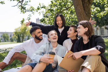 Excited asian student looking at cheerful interracial friends on grass in park  