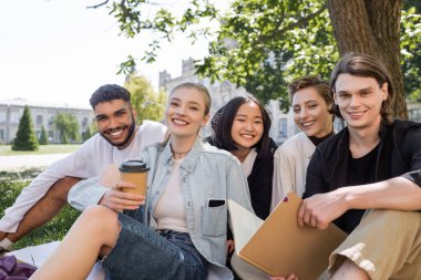 Positive multicultural students with notebook and coffee looking at camera on grass in park 