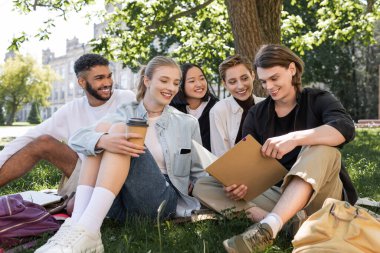 Smiling multicultural students looking at friend with notebook on grass in park 