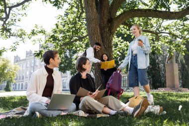 Student with coffee to go and backpack talking to interracial friends in summer park 