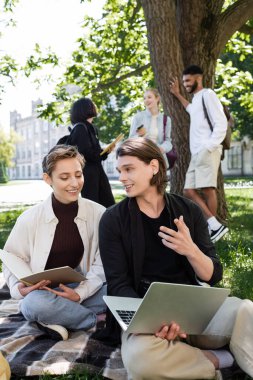 Student with laptop talking near friend with notebook on blanket in park 