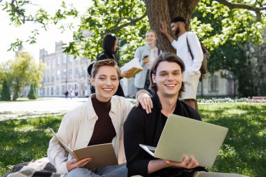 Smiling students with notebook and laptop looking at camera on grass in park 