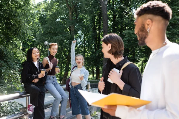 Excited multiethnic students holding coffee to go near blurred friends in park