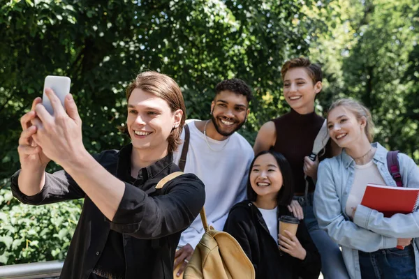 Uśmiechnięty Student Robi Selfie Smartfonie Pobliżu Międzyrasowych Przyjaciół Parku — Zdjęcie stockowe