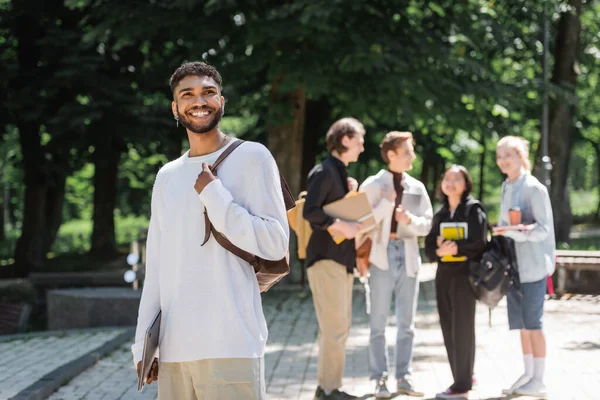 在夏天的公园里 拿着笔记本电脑站在模糊的朋友旁边笑着的非洲裔美国学生 — 图库照片