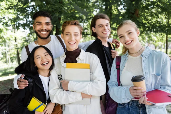 Cheerful interracial students with notebooks looking at camera in summer park