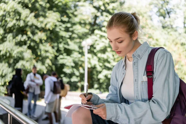 Student with backpack writing on notebook in park