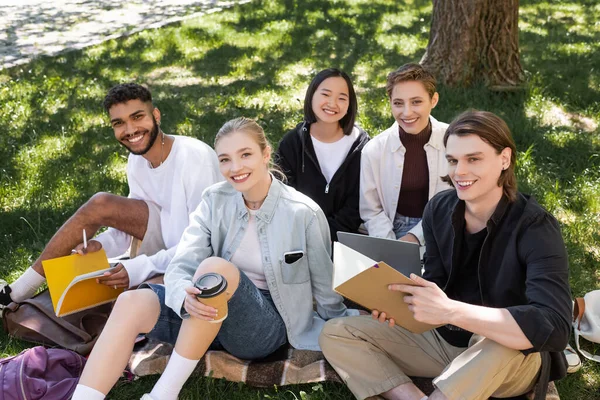 在公园里拿着笔记本和咖啡在草地上嬉笑的跨种族学生 — 图库照片