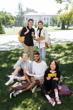 Cheerful multiethnic students with notebooks and coffee looking at camera on grass in park 