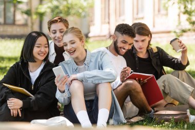 Smiling interracial students looking at smartphone near friends with notebook on lawn in park 