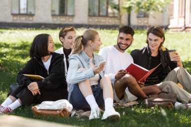 Multicultural students with smartphone and copy books spending time on grass in park 