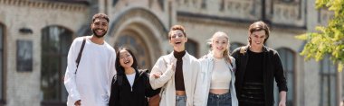 Positive multiethnic students looking at camera near blurred university outdoors, banner 