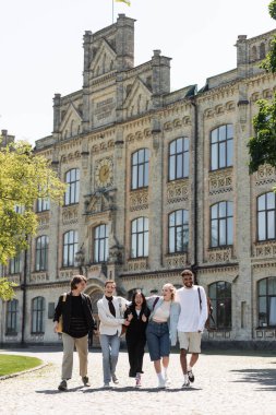 Sırt çantalı gülümseyen ırklar arası öğrenciler üniversitenin yakınlarında yürüyorlar. 