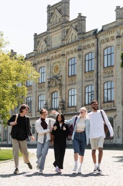 Positive multiethnic students with backpacks walking near blurred university outdoors 