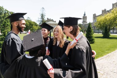 Smiling interracial bachelors in gowns holding diplomas and hugging in park 