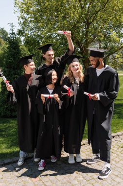 Smiling interracial bachelors looking at african american friend with diploma in park 