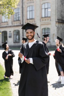 Önlüklü, genç, Afro-Amerikalı bekar diplomalı ve parktaki kameraya bakıyor. 