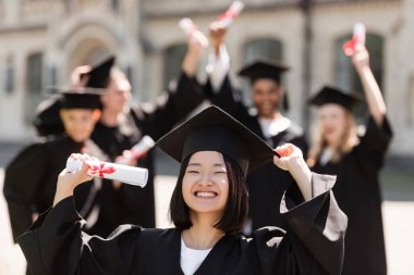 Şapkalı ve cüppeli gülümseyen Asyalı bekar açık havada bulanık arkadaşlarının yanında diploma taşıyor. 