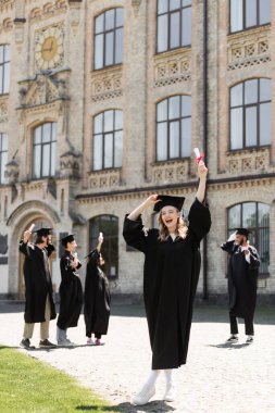 Excited bachelor holding diploma near blurred multiethnic friends and university outdoors 
