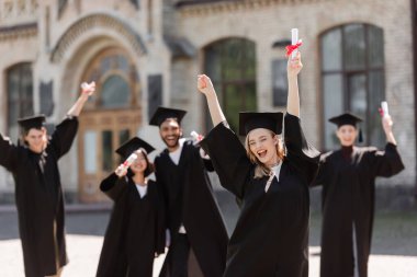 Excited student in bachelor gown holding diploma near multiethnic friends outdoors 