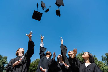 Interracial bachelors with diplomas throwing caps with sky at background 