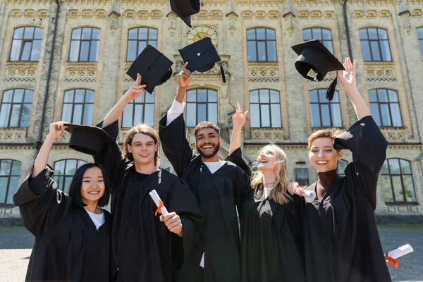 Smiling Multiethnic Bachelors Holding Diplomas Throwing Caps Outdoors — Stock fotografie